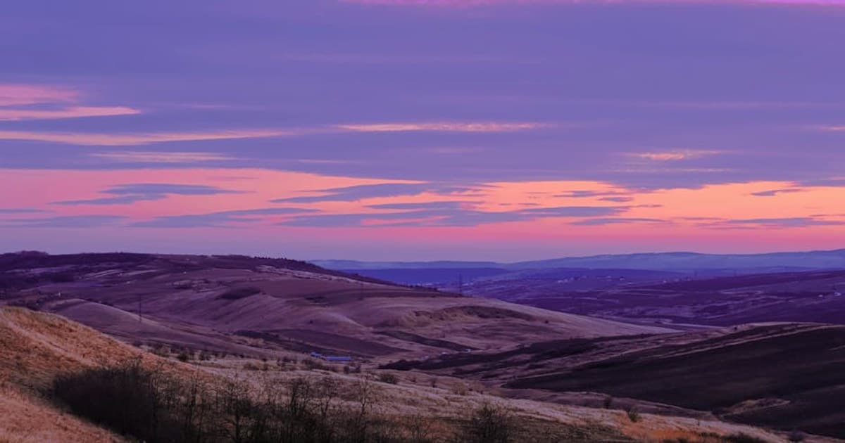A stunning sunrise paints the sky over rolling hills with serene hues of purple and pink.