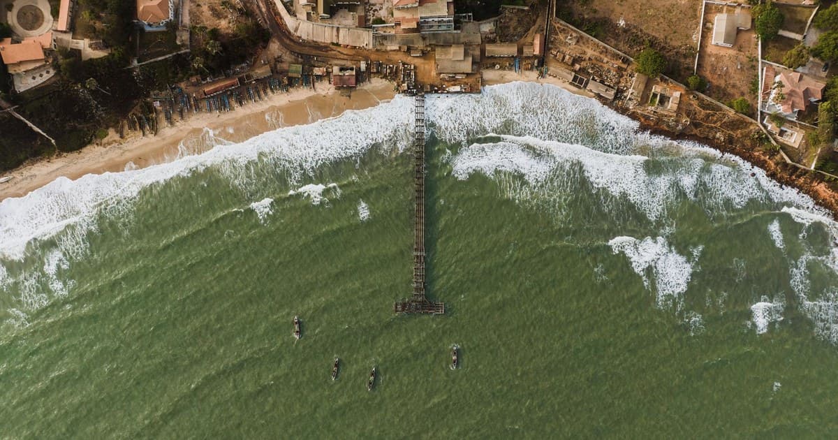 Drone shot of a coastal village with a pier, boats, and crashing waves.